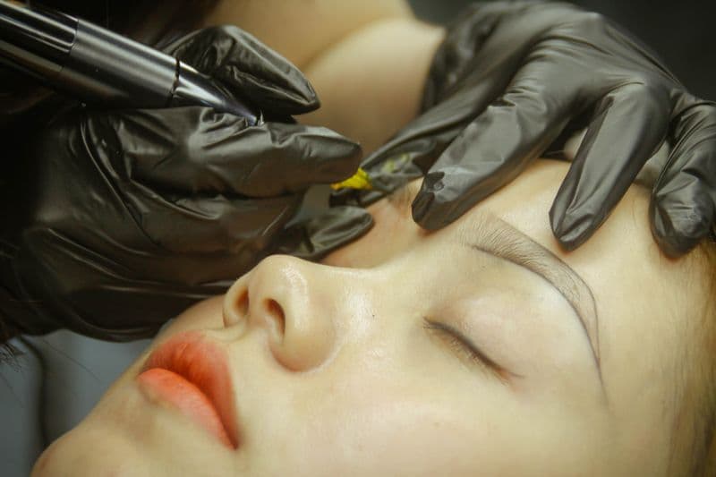 Microblading eyebrow procedure with gloved artist applying hair strokes — healing aftercare guide